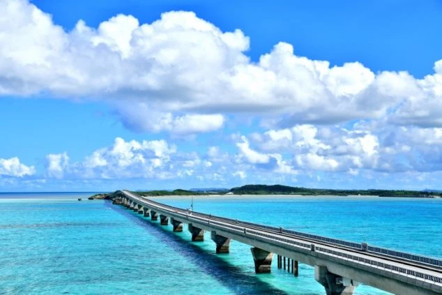 ターコイズブルーの海の上に架かる長い橋の全景。空は青く、もくもくと大きな白い雲が浮かんでいる。橋は遠くの島へと続いている。