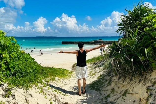 砂浜の小道から、青い海と白い砂浜を見ている男性の後ろ姿。男性は両腕を広げており、空は青く、白い雲が浮かんでいる。小道の両脇には緑の植物が生い茂っている。