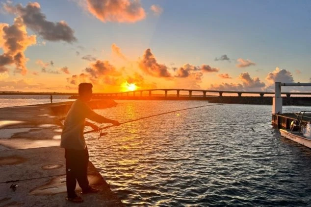 夕焼け空を背景に、防波堤の上で釣りを楽しむ男性のシルエット。夕日が水面に反射し、遠景には橋のシルエットが見えている。