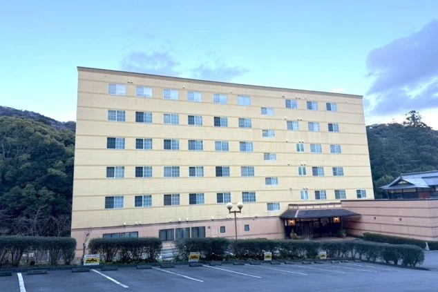 山あいに建つ、クリーム色の多層階ホテル。屋外駐車場とエントランスが見える。