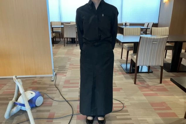 レストランの制服を着た人物。黒いシャツと黒いロングエプロンを着用しており、部屋の隅には掃除機が置かれている。