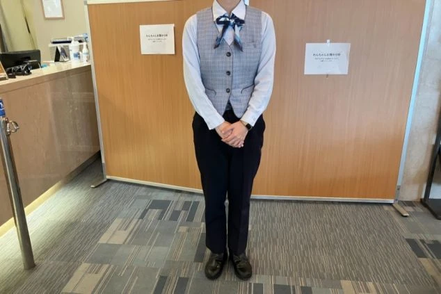 フロントの制服を着た人物。白いシャツにチェック柄のベスト、蝶ネクタイ、そして黒いズボンを着用している。