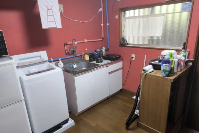 リゾートバイトの寮の共用キッチンの画像。赤い壁が特徴的な部屋に、洗濯機と冷蔵庫、シンクが備え付けられている。リゾートバイトの寮の共用キッチンの画像。