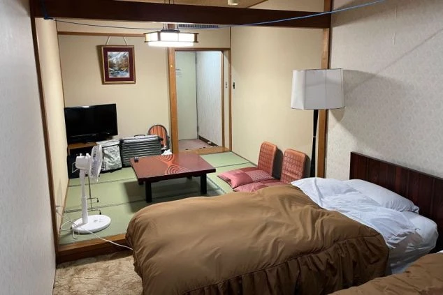 和室と洋室が一体となったリゾートバイトの寮の部屋。ベッド、テーブル、テレビ、扇風機が備え付けられている。