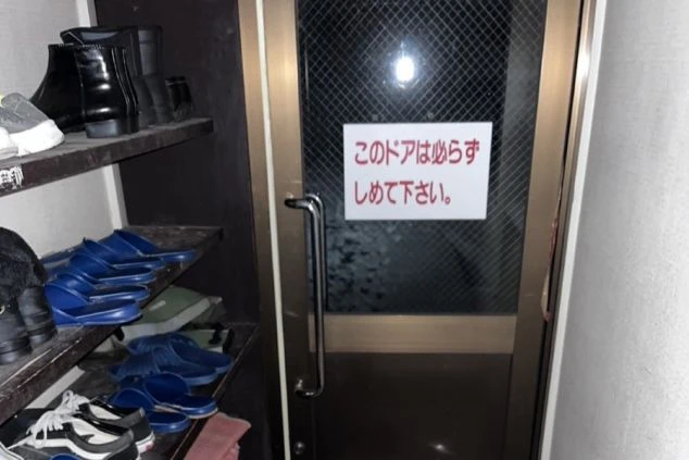 入り口のドアと靴箱の画像。棚には靴が並び、ドアには「このドアは必ずしめて下さい。」という注意書きがある。リゾートバイトの寮の共用玄関。