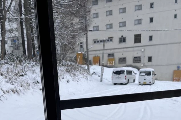 雪が積もった寮の屋外の風景の画像。建物の前に2台の白い車が停まり、窓のフレーム越しに見える。