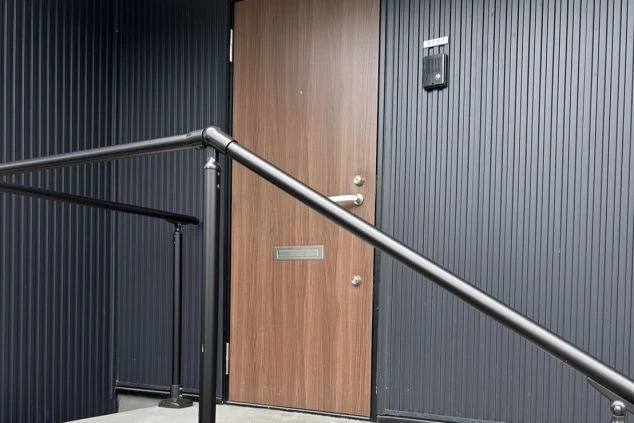  黒い波型の外壁と木製のドアがある建物の外観。手前には階段と手すりが見える。