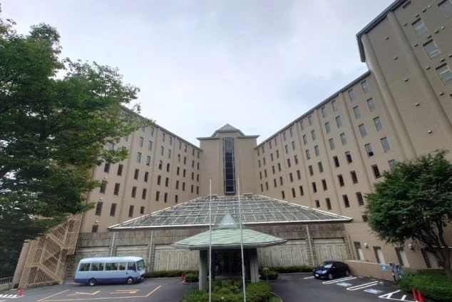 ベージュ色の壁の左右対称な高層棟と、中央にガラス張りの大きな屋根を持つエントランス（車寄せ）が見える。手前には送迎バスが停まっている。