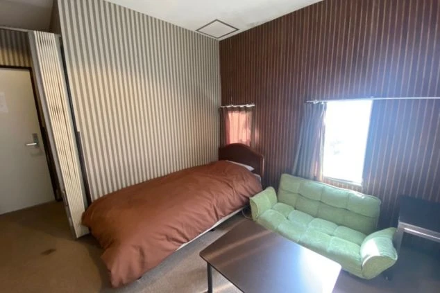 ベッド、ローテーブル、緑のソファが置かれたリゾートバイトの寮の個室の画像。