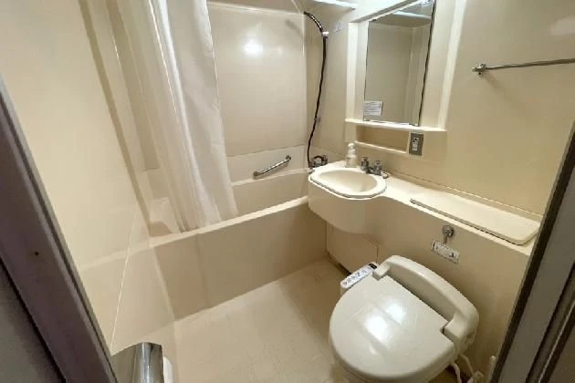 清潔感のあるコンパクトなユニットバス。白いシャワーカーテンが引かれ、洗面台の鏡が正面に見える。