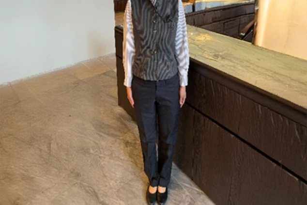 リゾートホテルのフロント(女性)制服。ストライプのシャツと黒のベスト、パンツスタイルで、接客業にふさわしいきちんとした服装。