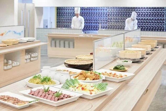 清潔感のある広々としたビュッフェ会場。手前にはローストビーフやサラダなどの料理が並び、奥のオープンキッチンでは白いコック帽を被った料理人が調理をしている。