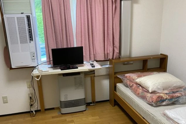 寮の個室。ベッド、机、テレビ、窓用エアコン、暖房器具が映る部屋の様子。