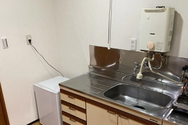 寮のキッチン。シンク、冷蔵庫、壁掛け給湯器が設置されたコンパクトな空間が映る。