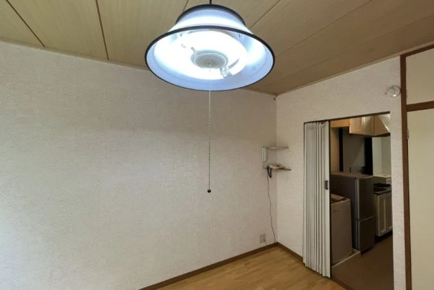 ワクトリの社員が出張で撮影した、リゾートバイトの寮の部屋の画像。フローリングの床と白い壁のシンプルな空間に、天井から照明が吊るされている。奥にはキッチンが見え、生活に必要な設備が整っていることを示す。