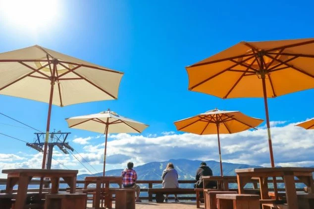 パラソルが広がるテラス席に座り、遠くの山々を眺める3人の人物の後ろ姿が写っている画像。晴れた青空が広がり、リゾートバイトの休日に岐阜県の鷲ヶ岳高原で雄大な自然を眺めながらゆったりと過ごすイメージ