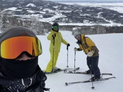  雪の中でスキーウェアを着た3人が写っている。一人はスキー板を履いており、もう一人はかがんでいる。