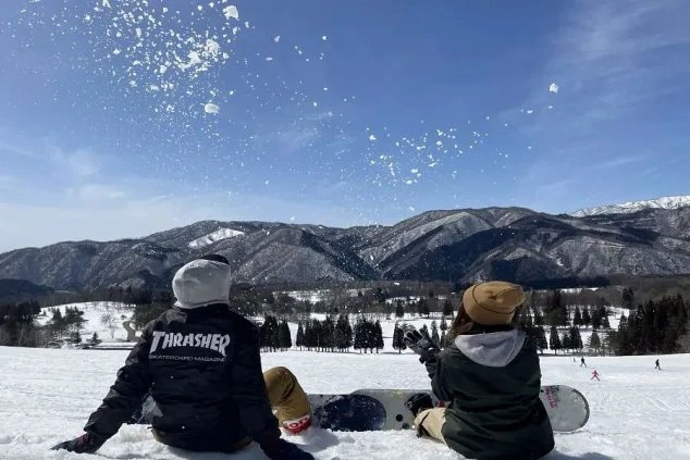雪山を背景にスノーボードを持って座り、景色を楽しむリゾバ仲間。リゾートバイトで出会った友人と過ごす、最高の休日のワンシーン。 選定のポイント：