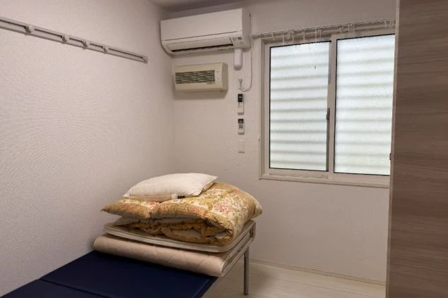 採光のよい個室(居室)。エアコンと暖房機が壁に取り付けられ、窓際には布団が積まれている。