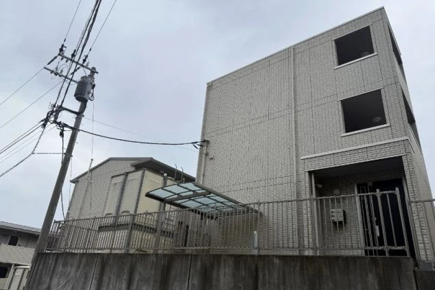 従業員寮の正面。灰色のタイル壁の三階建ての建物で、階段を上がったところに玄関扉がある。