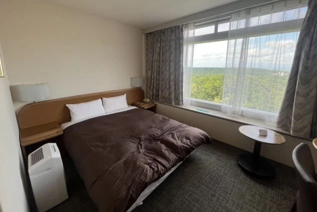 リゾートバイトの寮の部屋の中を撮影した画像。部屋にはベッド、デスク、ドレッサーが備え付けられており、窓からは豊かな自然が見える。リゾートバイトの寮の部屋の画像。