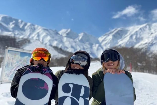晴れたスキー場でスノーボードを抱えて笑顔を見せる3人の男女。雪山を背景に、冬のリゾートバイトでウィンタースポーツを満喫できる楽しさが伝わるワクトリオリジナル画像。