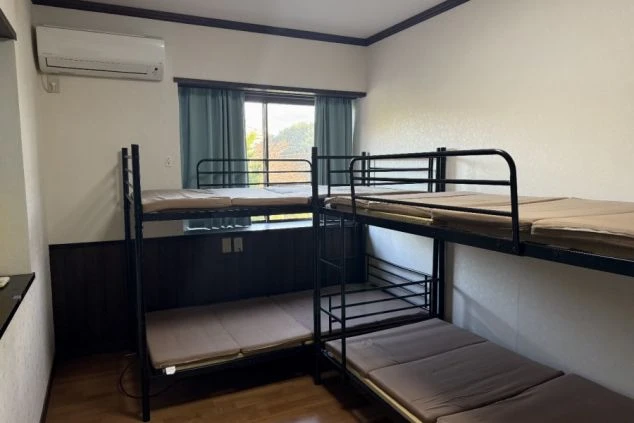 寮の個室内に並んだ黒いスチール製の2段ベッド。エアコンが完備され、窓にはブルーのカーテンがかかっています。