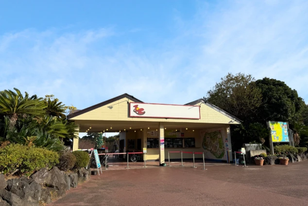 青空の下に建つ伊豆ぐらんぱる公園のチケット売り場。黄色の平屋建ての建物で、周囲にはヤシの木などの豊かな植栽があります。