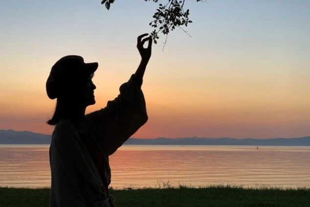 夕焼けの湖畔に立つ女性のシルエット。手を伸ばし、空と水面が染まる絶景が映るイメージ画像。リゾートバイトの休日を満喫。