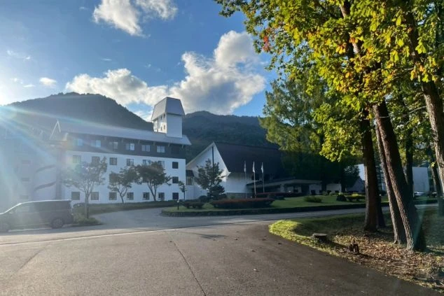 山々に囲まれたリゾートホテルまたはロッジの外観。白を基調とした建物で、左側にはチャペルのような尖った屋根が見える。青空と緑の木々が写り、太陽の光が差し込んでいる。