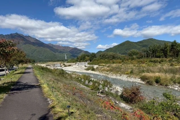 長野県大町市から望む、北アルプスの雄大な山々と清流の画像。遊歩道が続くのどかな田舎道から、冠雪した連峰と青空が広がる絶景。