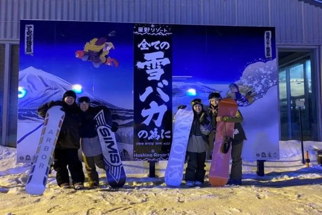リゾートバイト仲間が、スキー場の看板前でスノーボードを持って記念撮影。冬のリゾート地で、仕事も遊びも充実した毎日。