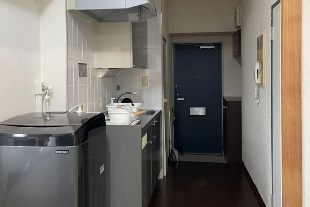 玄関と廊下が写されていて中央にはレンジフードのあるコンロとシンクエリアが見えます。カウンタートップは暗い色で、その下には引き出しや戸棚があるように見えます。