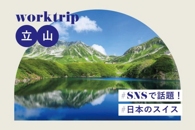 青空と残雪の山を背景に、緑の草原の中を石畳の道が真っ直ぐ続く風景。道の先には木造の低い建物が建っている。