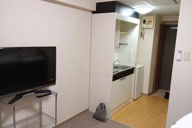 寮の居室全体。窓にはカーテンがかかり、暖房器具とテレビが設置されている。奥には簡易的なキッチンが見える。