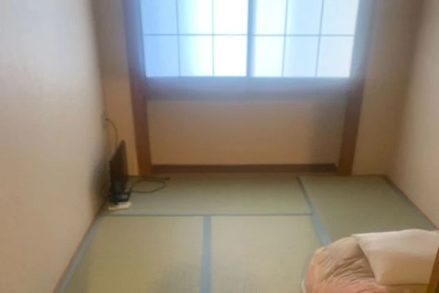  床にタタミが敷かれ、壁が薄いピンク色の和室の画像。部屋の奥には大きな窓があり、外からの光が差し込んでいます。窓の下には何か家具のようなものが置かれています。