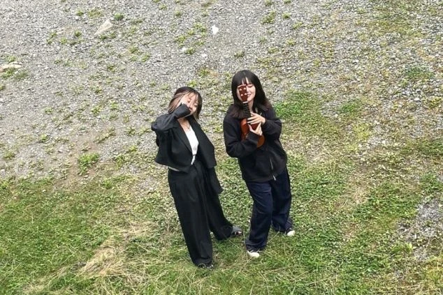 俯瞰で撮影された、白い花と緑の葉が広がる野原に立つ2人の人物の画像。2人は黒い服装をしており、カメラに向かってポーズをとっているように見えます。