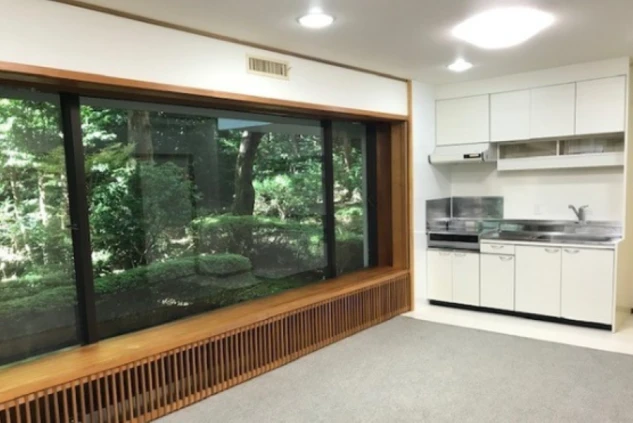 リゾートバイトの寮の、大きな窓から緑豊かな庭が望めるキッチン付きの広々とした部屋。窓際には木製の長いベンチが備え付けられている。