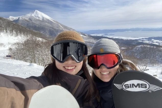 スキー場でスノーボードを楽しむ人々の自撮り画像。ゴーグルを着用し笑顔で、遠くには雪山と湖が見える。リゾートバイトの休日にウィンタースポーツを満喫している様子。