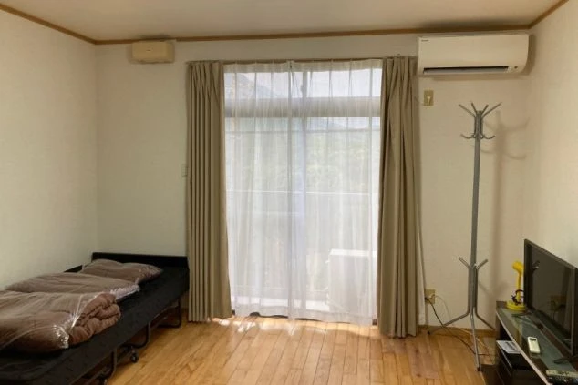 リゾートバイトの寮の部屋の画像。フローリングの床にベッド、テレビ、エアコン、コートハンガーが備わっている。