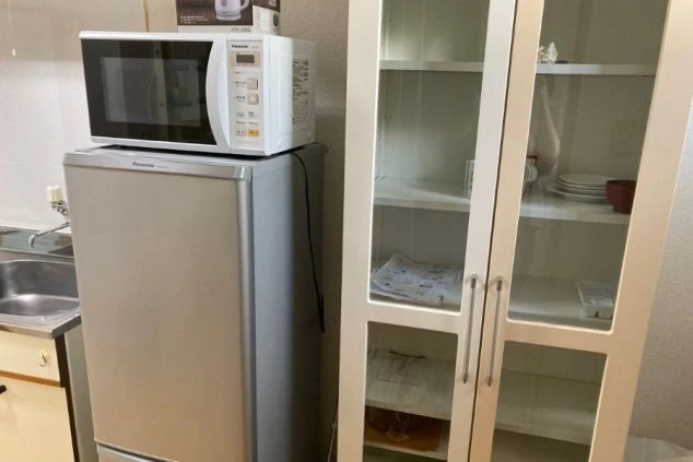 リゾートバイトの寮のキッチンの画像。冷蔵庫の上に電子レンジと電気ケトルが置かれ、食器棚も見える。自炊に便利な設備。