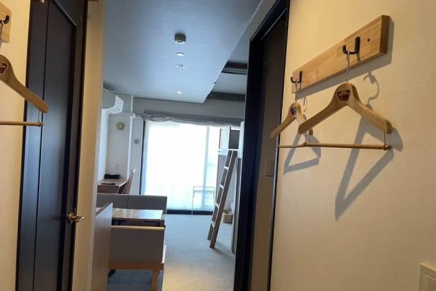 部屋の入り口から見た寮の様子。手前にはハンガーが掛かったフックがあり、奥にはベッドやテーブルが見える。