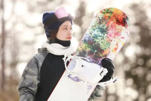 リゾートバイトの休日に、スノーボードを持って笑顔を見せる女性。冬のゲレンデで、仕事の合間にリフレッシュする様子。