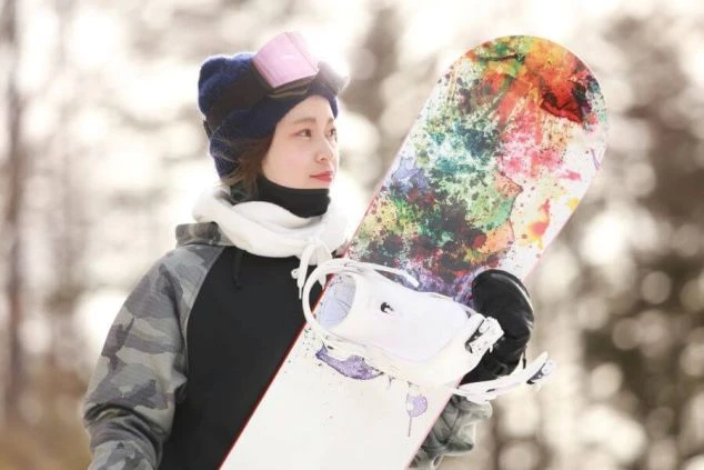 カラフルなスノーボードを肩にかけた、スキーウェア姿の女性。冬の澄んだ空気の中で、凛とした表情を見せています。