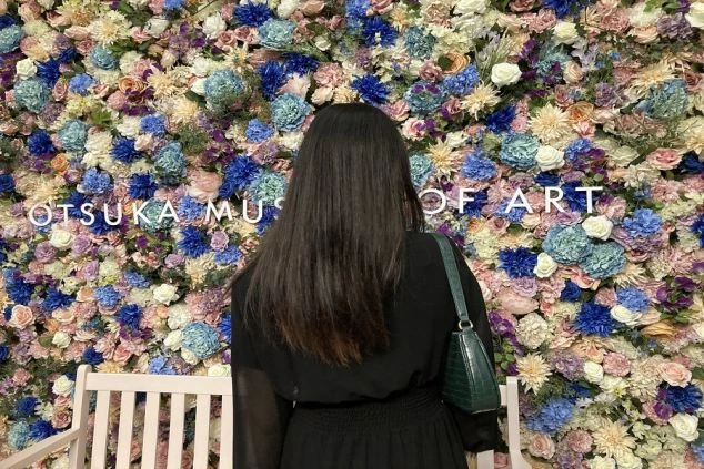 青、紫、白など様々な色の造花で覆われた壁の前に立つ、黒い服を着た女性の後ろ姿。