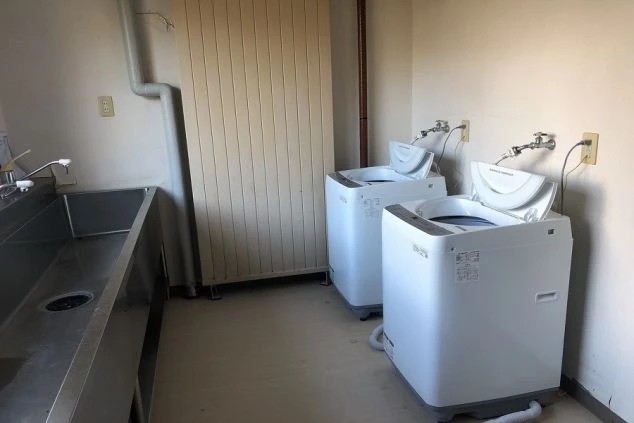 寮内にある共有のランドリールームの画像。全自動洗濯機が複数台設置され、横には広々としたシンク（洗い場）も完備。