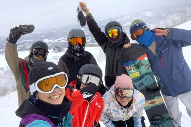 ワクトリでリゾートバイトをしたスタッフが撮影した、雪山でカメラに向かって笑顔でポーズをとる、ヘルメットやゴーグルをつけた6人のスノーボーダー。一人がスノーボードを持っている。