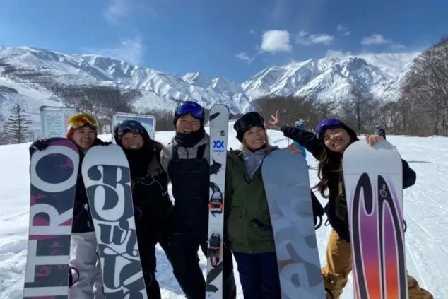 雪山を背景にスノーボードを持って笑顔でポーズを取るリゾートバイト仲間。冬のリゾート地で、仕事も遊びも充実した毎日。