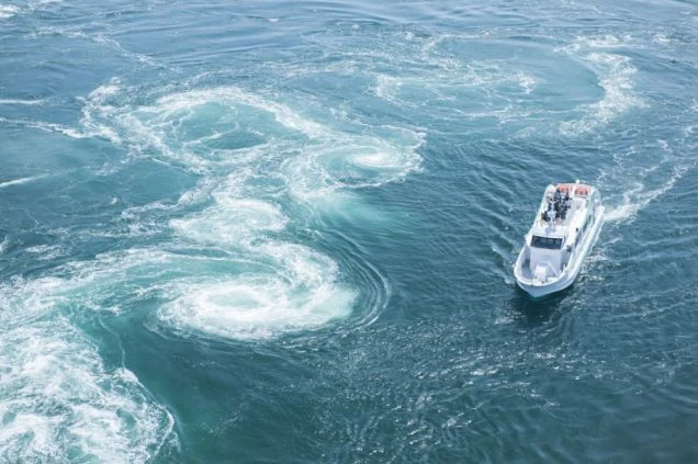 渦潮が巻く青緑色の海の上を航行する小さな観光船の俯瞰写真。船の周囲には、潮の流れによって発生した大きな渦巻きがいくつも確認できる。