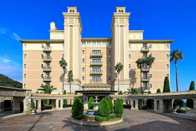 晴れた青空を背景に建つ、ヨーロッパの宮殿のような豪華なリゾートホテルの外観。正面には噴水のあるロータリーがあり、建物はツインタワーのような構造で、ヤシの木が植えられている。